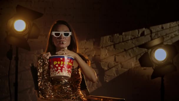 电影摄影和电影设置后台广告的概念 身穿金色衣服 戴着3D眼镜的女人坐在导演的椅子上 看电影 吃爆米花 垂下水桶 慢动作 — 图库视频影像