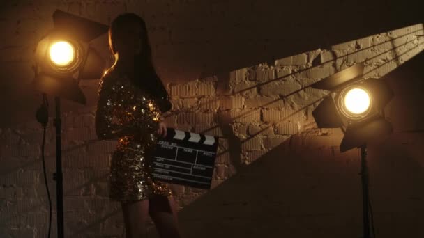 シネマトグラフィーと映画は舞台広告コンセプトを設定しました 白いレンガの壁の近くに立っている黄金のドレスのエレガントなブルネットの女性は クラッパーボードを保持するカメラにポーズ スローモーション — ストック動画