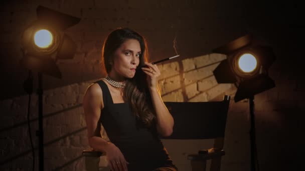 电影摄影和电影设置后台广告的概念 穿着黑色衣服的布鲁内特女人们坐在导演的椅子上 拿着烟盒抽烟 望着别处 微笑着 慢动作 — 图库视频影像
