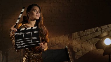 Sinematografi ve sinema sahne arkası reklam yaratıcı konsepti. Altın elbiseli güzel bir kadının, yönetmen koltuğunun yanında el çırpma tahtası tutarken ve kameraya bakarken yakın plan çekimi..