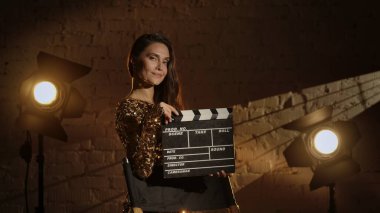 Sinematografi ve sinema sahne arkası reklam yaratıcı konsepti. Altın elbiseli çekici bir kadının, yönetmen koltuğunun yanında el çırpma tahtası tutarken ve kameraya gülümserken yakın çekimi..