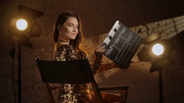 Sinematografi ve sinema sahne arkası reklam yaratıcı konsepti. Altın elbiseli esmer bir kadının yakın plan çekimi, yönetmen koltuğunda oturuyor, elinde alçıpan var ve kameraya gülümsüyor..