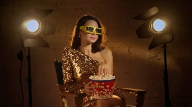 Sinematografi ve sinema sahne arkası reklam yaratıcı konsepti. Altın elbiseli esmer bir kadının yakın plan resmi, yönetmen koltuğunda oturuyor, kağıt bardaklar içinde ve kovadan patlamış mısır yiyor..