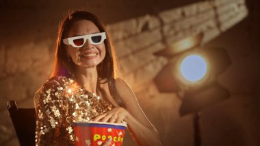 Sinematografi ve sahne arkasında film. Altın elbiseli güzel esmer kadın aktrisin yakın plan çekimi yönetmen koltuğunda oturup komedi izlerken 3D gözlük takıp patlamış mısır yerken..