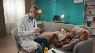 Hastanın evdeki durumunu kontrol eden bir doktorun tam boy videosu. Sağlık sorunları olan yaşlı bir adam tarafından tanımlanan belirtileri not etmek için tablet kullanıyor. Tıbbi hizmet, reklam..