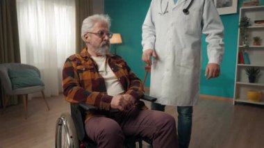Tekerlekli sandalyedeki engelli yaşlı bir hastanın evde sağlık kontrolü yaptığı orta büyüklükte bir video. Doktor omzuna hafifçe vuruyor. Sağlık sistemi, sağlık hizmetleri, reklam.