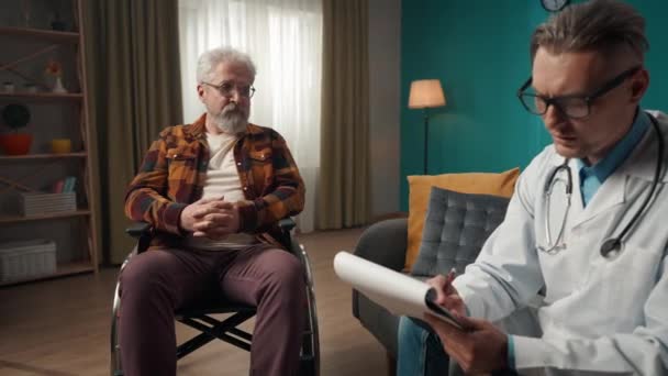 一个医生在家中检查病人病情的全片录像 他正在和一个坐轮椅的老人说话 在记事本上写下一些东西 — 图库视频影像