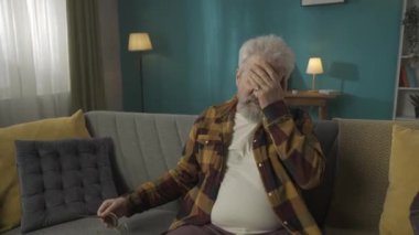 Yaşlı, emekli bir adamın orta ölçekli videosu, koltukta oturan yaşlı bir vatandaş, şiddetli baş ağrısı nedeniyle boynuna ve kafasına masaj yapıyor, migren. Sağlık sistemi, sağlık hizmeti, reklam. HDR