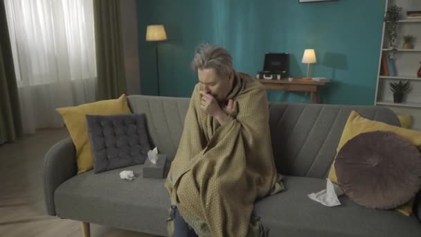 放大了一个中年男人坐在沙发上的视频 沙发裹着毛毯 咳嗽得好像他得了流感或感冒 他的流鼻涕用了很多纸巾 医疗保健 医疗服务 — 图库视频影像