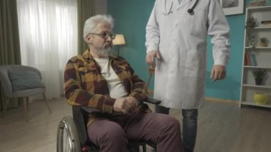 Tekerlekli sandalyedeki engelli yaşlı bir hastanın evde sağlık kontrolü yaptığı orta büyüklükte bir video. Doktor omzuna hafifçe vuruyor. Sağlık sistemi, sağlık hizmeti, reklam. HDR BT2020