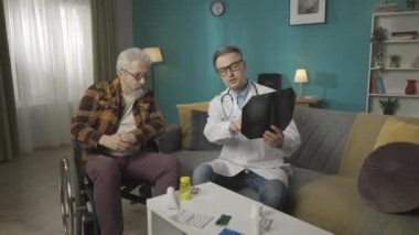 Hastanın evdeki durumunu kontrol eden bir doktorun tam boy videosu. Tekerlekli sandalyedeki yaşlı bir adamla konuşuyor, röntgen filmi gösteriyor, konuşuyor. Sağlık sigortası, sağlık sigortası, reklam. HDR