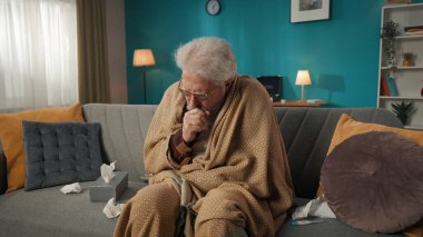 Yaşlı, emekli bir adamın orta boy fotoğrafı, koltukta oturan yaşlı bir vatandaş, battaniyeye sarılı bir kanepe, öksürük, kullanılmış mendillerle çevrili. Sağlık hizmetleri, sağlık sorunları, hastalık, reklam.