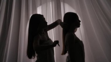 Havluya sarılmış iki kız kardeşin, genç kadın siluetinin orta ölçekli videosu. Biri okşuyor, diğerinin saçlarını boğuk bir ışıkta tarıyor. Ürün reklamı, bakım rutini.