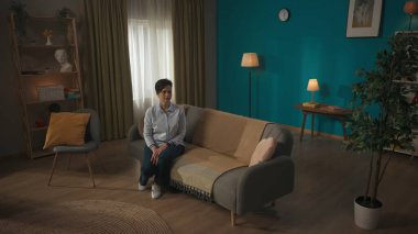 Orta yaşlı, kısa saçlı esmer bir kadının bir kanepede otururken, güneşli, rahat bir odada koltukta otururken, çerçevenin dışından birine ya da bir şeye bakarken fotoğrafı. İç mekan, iç mekan...