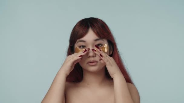 一个有着红色染发和裸体妆容的年轻的神童女人在她的眼皮底下涂上了修饰和拍拍眼罩的特写视频 黑眼圈美容 化妆品 护肤广告 — 图库视频影像