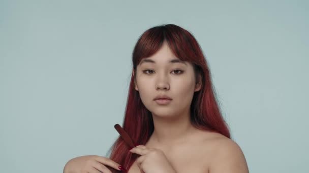 一个有着红色染发和裸体妆容的年轻女性凝视着相机 梳理着她柔滑的秀发的特写视频 化妆品 护发品广告 — 图库视频影像