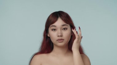 Kırmızı boyalı saçlı ve çıplak makyajlı genç bir kadının kameraya bakıp ucuz yüzüne yüz kremi sürdüğü yakın çekim yavaş çekim videosu. Güzellik, kozmetik, cilt bakımı.