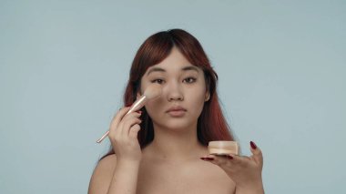 Kırmızı boyalı saçlı ve çıplak makyajlı genç bir kadının kameraya bakıp yüzüne pudra sürerken çekilmiş yakın plan fotoğrafı. Güzellik, kozmetik, cilt bakımı.