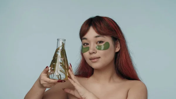 一个有着红色染发和裸体妆容的年轻的精液女子穿着眼罩的近照 照片上有一个装有海藻的实验室瓶子 黑眼圈美容 化妆品 护肤广告 — 图库照片