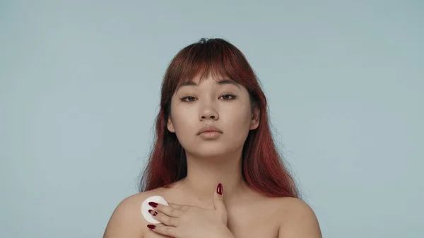 一个有着红色染发和裸体妆容的年轻女性在镜头前凝视着镜头 在她的锁骨上移动着一块棉垫 这是她的近照 护肤广告 — 图库照片