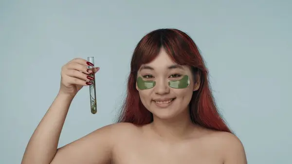 一个有着红色染发和裸体妆容的年轻的精液女子穿着眼罩的近照 照片上有一个装有海藻的实验室瓶子 黑眼圈美容 化妆品 护肤广告 — 图库照片