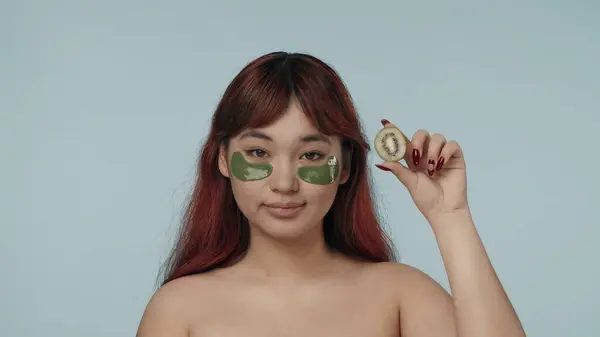 一个有着红色染发和裸体妆容的年轻女性穿着眼罩 看着相机 拿着一条剪下来的猕猴桃的特写照片 化妆品 护肤日常广告 — 图库照片