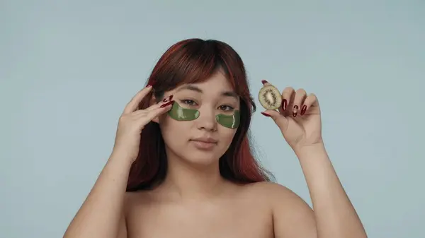 一个有着红色染发和裸体妆容的年轻女子 戴着眼罩 轻轻触摸着她们 拿着一个剪短的猕猴桃的特写照片 化妆品 护肤日常广告 — 图库照片