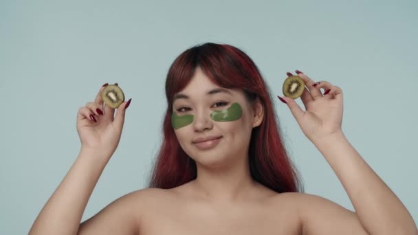 赤い染められた髪とヌードのメイクを施した若いセミナーの女性のクローズアップされた隔離されたスローモーションビデオは 彼女の目の前に2つのキウイを保持しています 美しさ 化粧品 スキンケア広告 — ストック動画