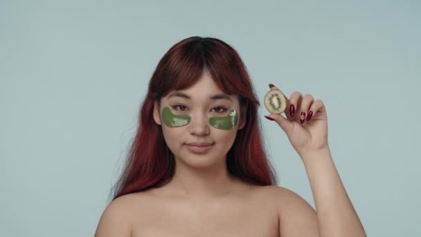 一个有着红色染发和裸体妆容的年轻女性穿着眼罩 轻轻拍拍她们 拿着一个剪短的猕猴桃的特写慢镜头 化妆品 护肤广告 — 图库视频影像