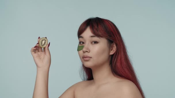 赤い染められた髪とヌードのメイクアップを施した若いセミナーの女性のクローズアップされたスローモーションビデオは カットキウイを保持し チェックに触れました 美しさ 化粧品 スキンケア広告 — ストック動画