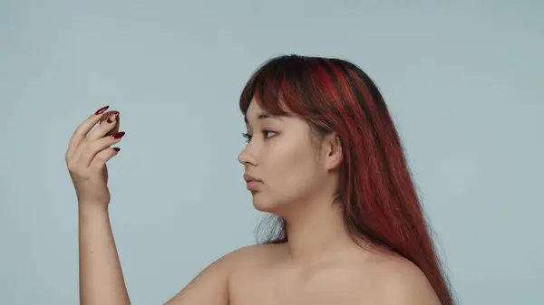 一个有着红色染发和裸体妆容的年轻的神童女子站在半圆形的特写镜头下 手里拿着一只猕猴桃 化妆品 护肤日常广告 — 图库照片