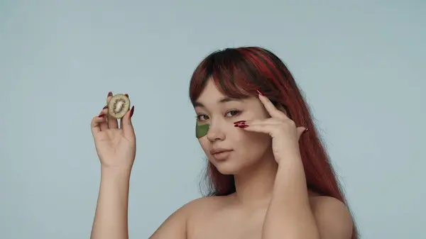 一个有着红色染发和裸体妆容的年轻女人 戴着眼罩 轻轻触摸着它 拿着一条剪下来的猕猴桃的特写照片 化妆品 护肤日常广告 — 图库照片