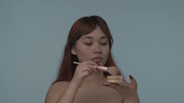 赤い染められた髪とヌードのメイクを施した若いセミナーの女性のクローズアップされたスローモーションビデオは 彼女の顔にパウダーを設定します 美しさ 化粧品 スキンケア広告 — ストック動画