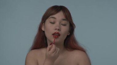 Kırmızı boyalı saçlı, çıplak makyajlı ve dudaklarını rujla boyayan genç bir kızın yakın çekim izole videosu. Güzellik, kozmetik, mua, ürün reklamı. HDR BT2020 HLG Malzemesi.