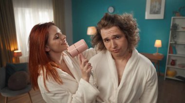 Genç bir çiftin yakın çekimi resmedildi. Canı sıkkın bir adam, yüzü aynada bakar gibi kameraya bakıyor. Yüzünde gülümseme olan kız saçını saç kurutma makinesiyle kurutuyor..