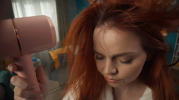 在一个有着红头发和化妆的女孩的特写中 闭上她的眼睛 她用粉红的吹风机擦干它们 然后往下看 头发在气流上生长 发型和发型 — 图库照片