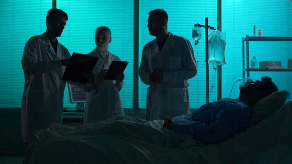 Full-sized silhouette shot of three doctors standing around a patient on a breathing support, in coma, laying in a hospital bed in front of them. Hospital, impatient treatment, medical care.