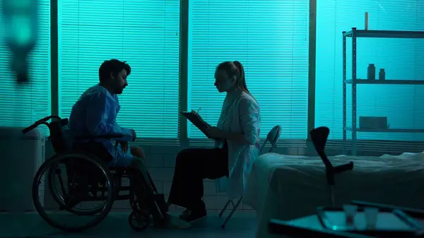 Full-sized silhouette shot a disabled man, patient with mobility impairment on a wheelchair getting a check-up. A female doctor, nurse asks him some questions. Hospital, rehabilitation, medical.