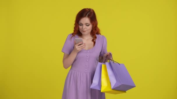 ショッピングバッグを持っている赤毛の女性は 期待している彼女のスマートフォンの画面に興奮しているように見えます 女性は喜んで叫び 勝利のジェスチャーを作ります 黄色の背景にあるスタジオの女性 セールス — ストック動画
