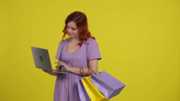 一个拿着购物袋的女人在用笔记本电脑 一个女人浏览利润丰厚的工作机会 促销活动 并在网上下订单 黄底画室里穿着浅色太阳衣的红头发女人 — 图库视频影像