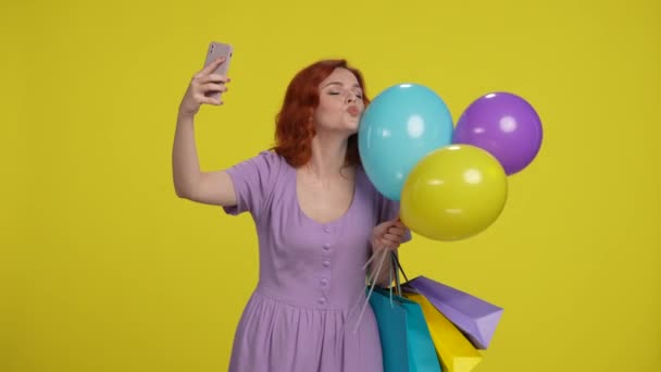 ショッピングバッグとカラフルな風船を持つ赤毛の女性は スマートフォンでセルフィーを取ります 黄色い背景で風船にキスをする女性 — ストック動画