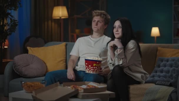 最初の愛と関係創造的なコンセプト 十代のカップルの肖像画 ソファーに座ってポップコーンを食べ 映画を見ている少年と少女 少年は恥ずかしがり屋の肩に手を置く 少女は抱擁を受け入れる — ストック動画
