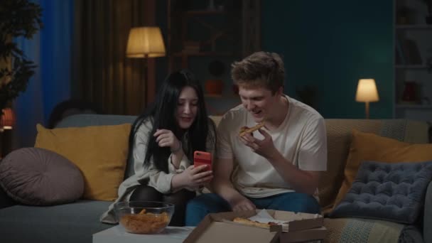 最初の愛と関係創造的なコンセプト 十代のカップルの肖像画は時間を費やしています ソファーに座っている男の子と女の子は ソーシャルメディアを見ているスマートフォンを保持し ピザを食べ セルフィー写真を撮る — ストック動画