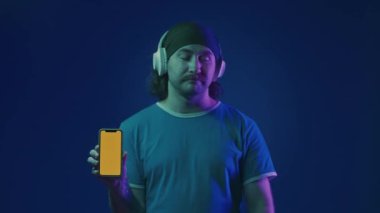 Beyaz kablosuz kulaklık takan bir adam işaret parmağını akıllı telefonunun yeşil ekranına doğrultuyor ve parmağını doğrultuyor. Stüdyoda kulaklığı ve akıllı telefonu olan mavi arka planda bir adam.