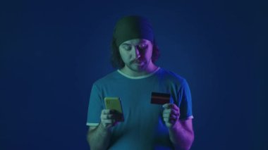 Çevrimiçi ödeme. Bir adam online bir mağazadan kredi kartı ve akıllı bir telefonla alışveriş yapıyor, onaylayarak başını sallıyor. Stüdyoda mavi arka planda yeşil ve pembe neon ışıklı bir adam.