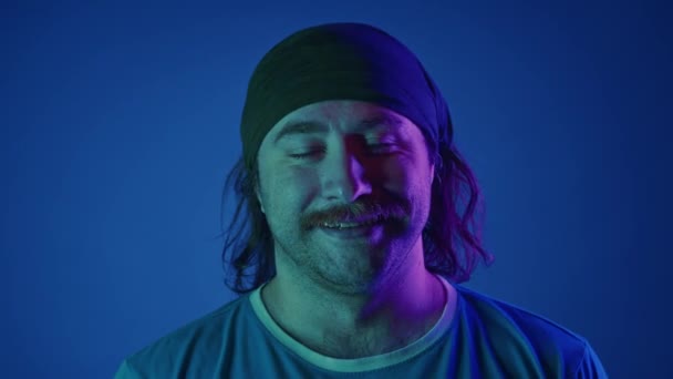 ヘッドバンドを閉じた笑顔のマッサージされた長髪の男性の肖像画 男はピンクと緑のネオンライトの青い背景に対してスタジオで深呼吸とエキゾールを取る — ストック動画