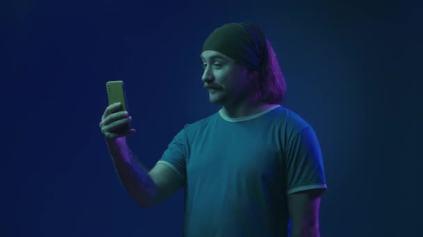 一个男人用智能手机在一个视频电话上说话 那个人挥挥手致意 在背后指指点点 说再见 粉红绿色霓虹灯蓝色背景的演播室里一个有胡须的男人 — 图库视频影像