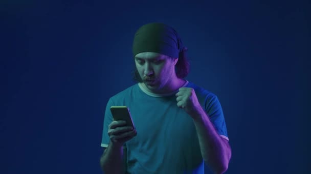 一个兴奋的男人紧张而期待地看着智能手机屏幕 男人欢呼雀跃 做了一个胜利的手势 一个拿着电话在工作室里拿着蓝色背景粉红的男人 — 图库视频影像