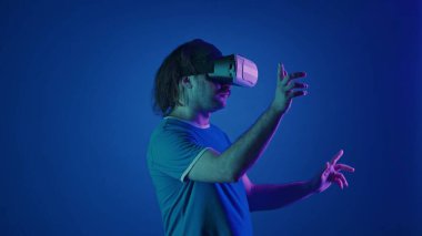 Sanal gözlük takan bir adam görünmez bir ekranı kaydırıyor. Stüdyoda, mavi arka planda, pembe ve yeşil neon ışıklı bir adam. Gelecekçi teknoloji
