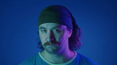 Bıyıklı bir adamın portresi stüdyoda, mavi arka planda, pembe ve yeşil neon ışıkta. Adam dümdüz bakıyor, dikkatlice dinliyor.